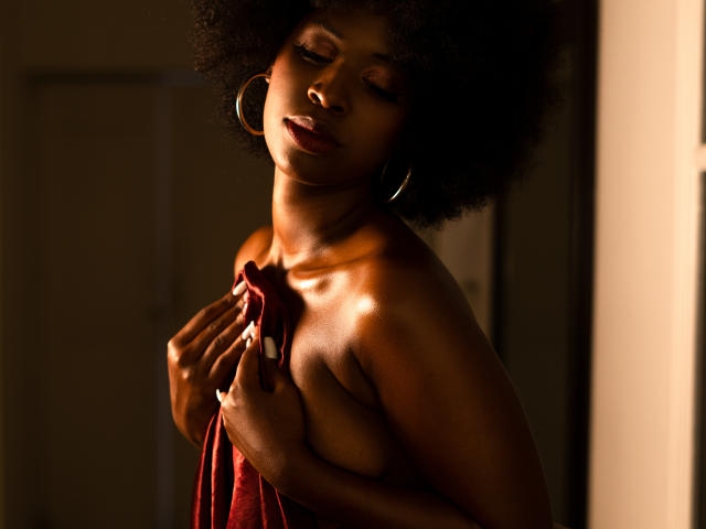 Karmen Castillo, met donkere huid en een afro, houdt een rode doek voor haar kleine borsten, draagt oorbellen met ringen, heeft haar ogen dicht en staat in zacht zonlicht. Haar nagels zijn lang en wit..