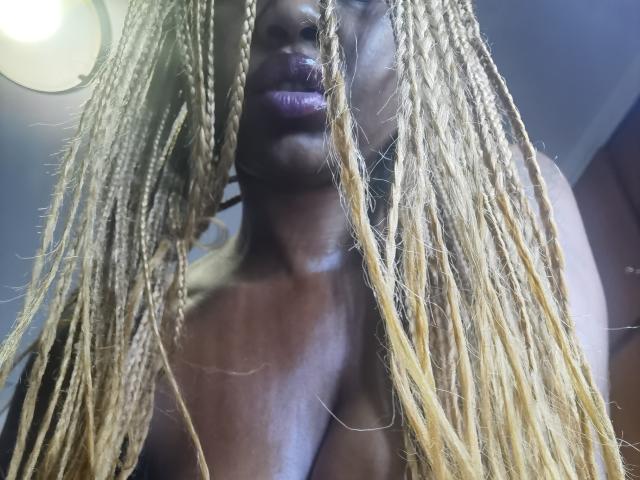 LaPicante with dark skin, purple lipstick, and long blonde braids covering her chest, looking down with a slight smile. Her tits are barely visible through her hair.