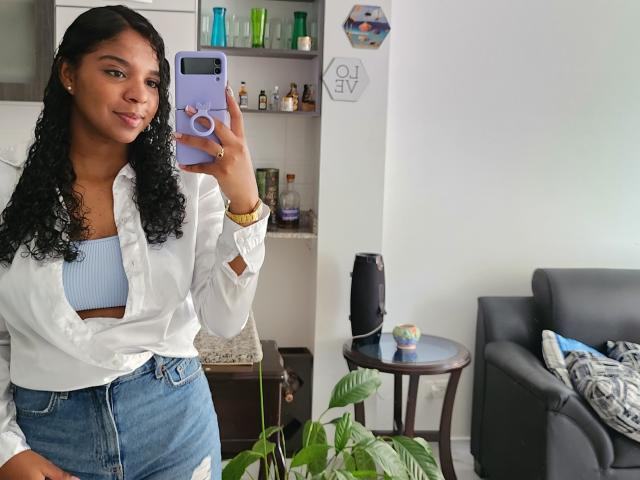 LisaMendez with curly black hair, light brown skin, wearing a white shirt, blue crop top, and high-waisted jeans, holding a purple phone, standing in a room, smiling.