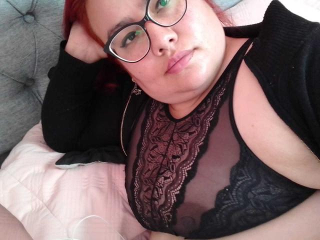 FoxyBell with red hair, glasses, black lace bra showing big tits, black zip-up, lying on pink bed, pouty face, right arm behind head, looking at camera.