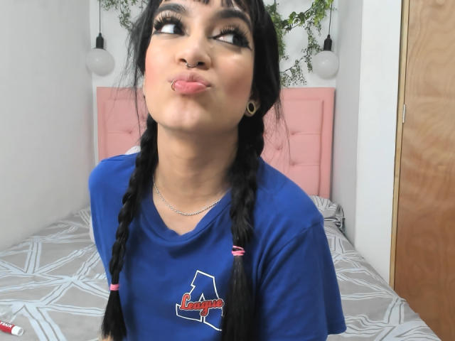MeliMuller with black braids, pouting lips, nose ring, blue shirt, and large eyelashes, puffy eyes, sitting on a bed with a pink headboard, looking up.