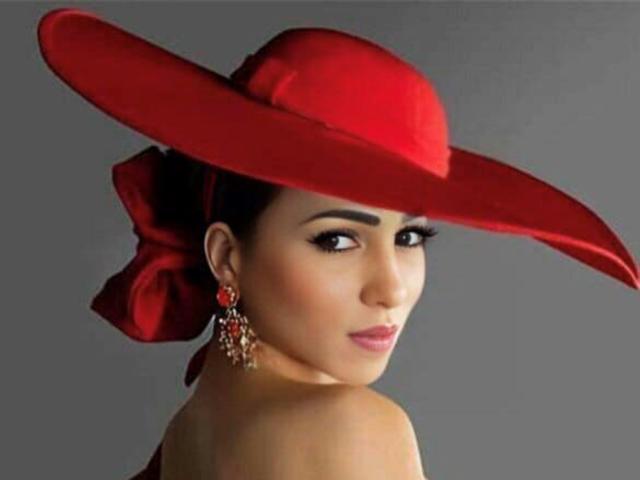 Vanna Garcia, con un cappello rosso, un'acconciatura a chignon con capelli rossi, orecchini rossi e spalle scoperte, guarda indietro. Ha un viso attraente e un sorriso malizioso. Il suo seno non è visibile..