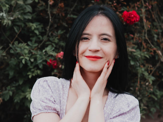 AlanaMoneth has black hair, red lipstick, and a light purple patterned shirt. She's smiling with hands on her cheeks, showing red nails. Her tits are small and her pussy is not visible.