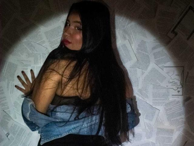 RossyGranger with long black hair, looking back over shoulder, wearing a blue denim jacket, exposing bare shoulders, hands on wall, dark red lipstick, slightly smoky eye.