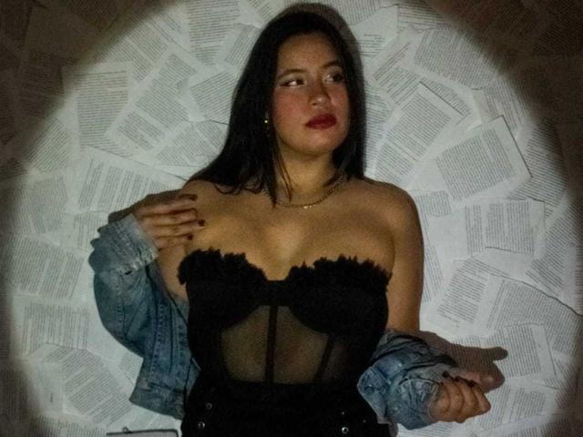 Rossy Granger draagt een zwarte, mouwloze korset, waarbij haar borsten zichtbaar zijn. Ze draagt een denimjasje, haar handen rusten op haar borst, ze heeft rode lippenstift op, donker haar en een gouden ketting. De foto is genomen in een omgeving met weinig licht..