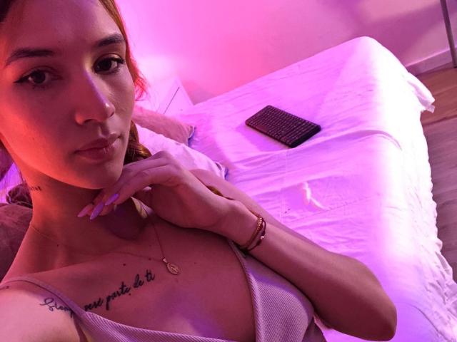 Greta Harris, com pele clara, cabelo rosa, seios pequenos, tatuagem no peito, vestindo um sutiã branco, quarto rosa-roxo, cama com lençóis brancos, controle remoto preto, pulseira, olhando para a câmera, expressão de lábios franzidos..