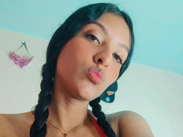 VickyHoney, avec ses tresses foncées, ses lèvres roses et pulpeuses, sa peau brune et un maquillage discret, porte un haut rouge et regarde en bas l'appareil photo. Un petit collier en or est visible..