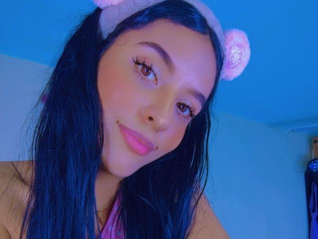 VickyHoney, com cabelos longos e pretos, uma faixa de cabeça rosa com orelhas de urso, lábios rosados, olhos castanhos e maquiagem discreta, sorrindo levemente em uma selfie com iluminação azul..