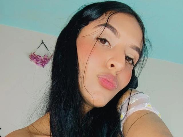 VickyHoney a de longs cheveux noirs, des yeux marron et des lèvres roses et pulpeuses. Elle porte un haut fleuri, regarde vers le bas avec un léger sourire, et porte un joli collier rose..