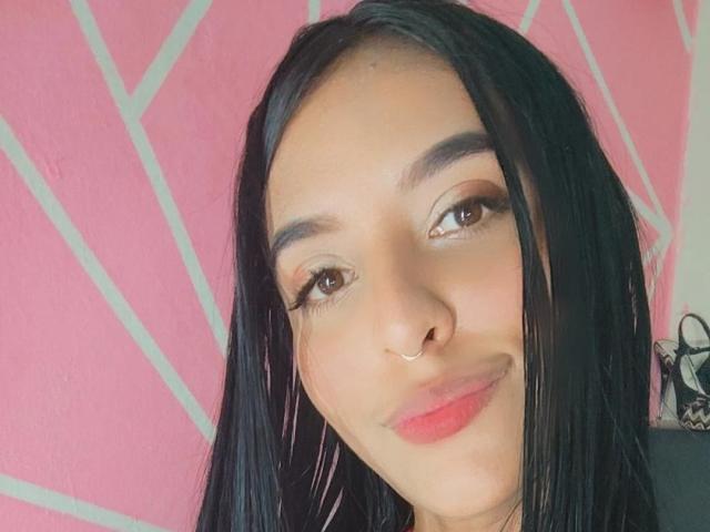 VickyHoney tem cabelo preto liso, olhos castanhos, um piercing no nariz e batom rosa. Ela está sorrindo com uma expressão adorável, olhando para a câmera. Seus seios não estão visíveis..