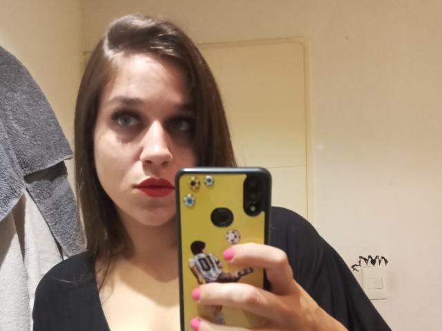 Ein Mädchen mit rotem Lippenstift, einem schwarzen Oberteil und rosafarbenen Nägeln, das ein gelbes Handy mit Aufklebern hält und ein Selfie vor einem Spiegel macht. Sie hat braunes Haar und einen neutralen Gesichtsausdruck..