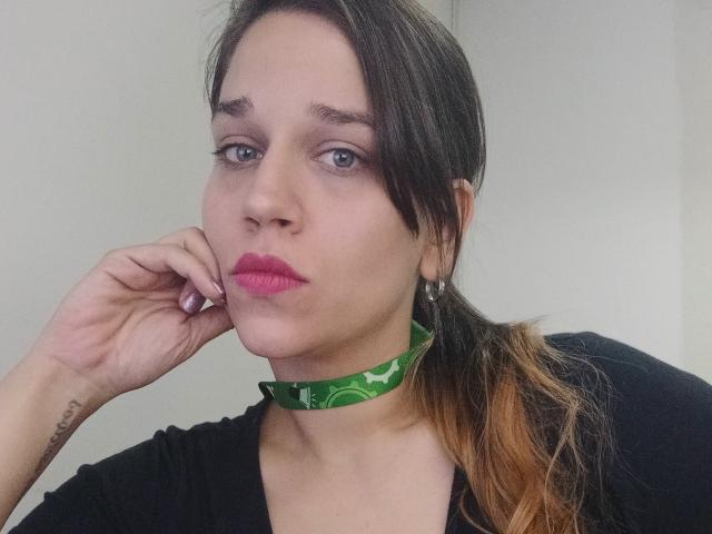 Ein Mädchen mit rosafarbenem Lippenstift, einem grünen Choker, einem schwarzen Hemd und einem braunen Pferdeschwanz, die ihr Kinn in die Hand legt und dabei ein Tattoo an ihrem linken Handgelenk zeigt..