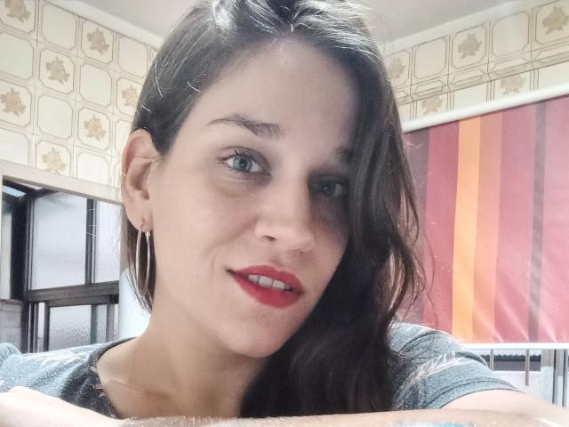 Faroles hat roten Lippenstift, braune, wellige Haare, blaue Augen und trägt goldene Ohrringe. Sie lächelt, hat die Arme verschränkt und trägt ein graues Hemd. Sie sieht süß aus..