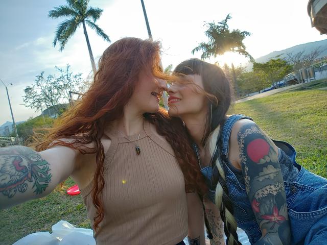 BethAndRoseI with red hair and tits in a ribbed tank top, and Rose with black hair and tattoos in denim, smiling and kissing in a sunny park.