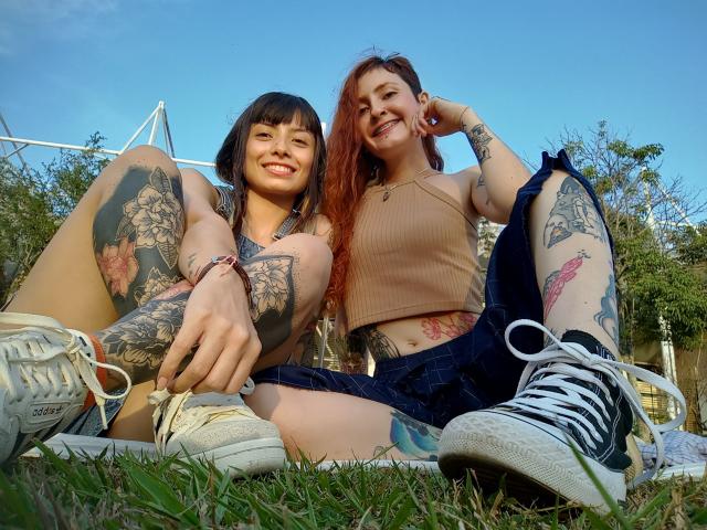 BethAndRoseI with tattoos, sitting on grass, wearing crop tops and shorts, smiling, white sneakers, blue sky, redhead with long hair, brunette with bangs, tattoos on arms and legs.