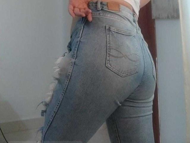 O traseiro de Sharon Greys está em calças jeans azuis apertadas e rasgadas, com bordas desfiadas, que mostram suas curvas e sua mão puxando a cintura. Sua blusa branca está parcialmente visível..