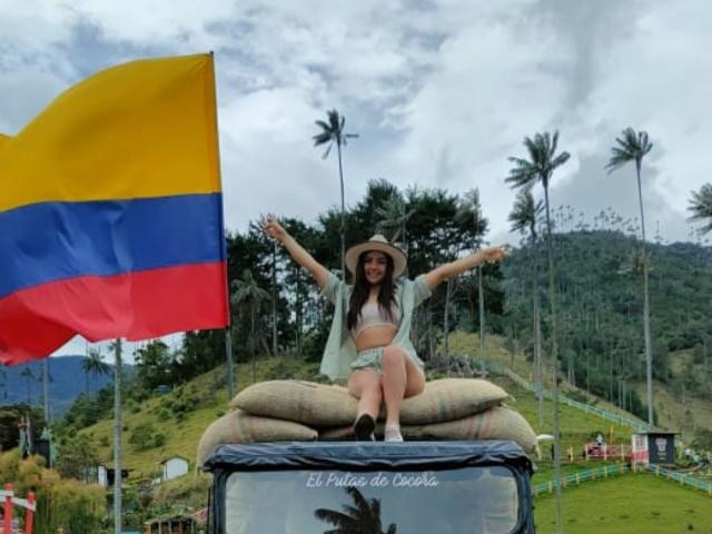 HeidyMillerr, zittend op jutezakken, met armen gespreid, een hoed op, een witte beha, een groene jas en een korte broek, voor een grote Colombiaanse vlag, met palmbomen op de achtergrond..