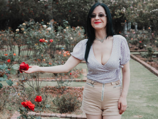 AlanaMoneth with black hair, red lipstick, glasses, cleavage, polka dot top, tan shorts, holding red rose, smiling, standing in garden, daytime.