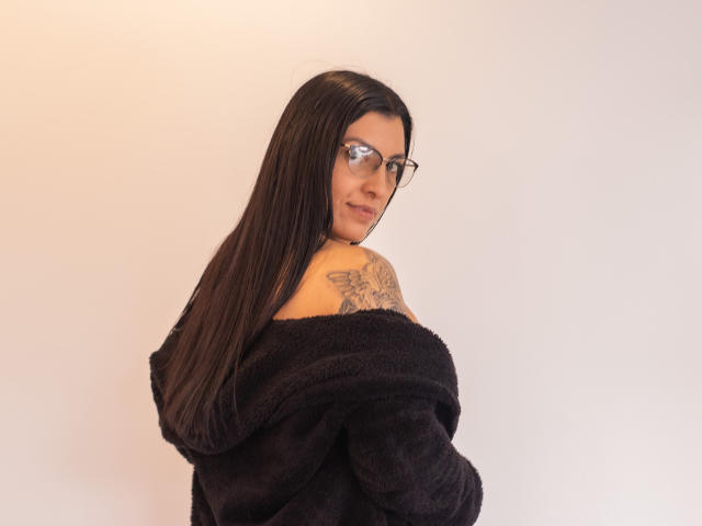 JanetteRider, avec de longs cheveux noirs, des lunettes, un pull noir douillet à épaules dénudées, un tatouage sur l'épaule droite, regardant en arrière, un léger sourire, peau claire, fond blanc, plan de la partie supérieure du corps..