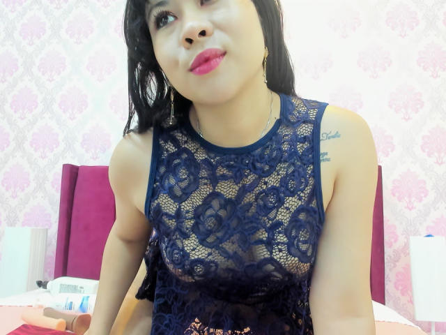 LourrieAnn with dark hair, wearing a sheer navy lace top, showing her small tits through the fabric, pink lipstick, and a tattoo on her right shoulder.