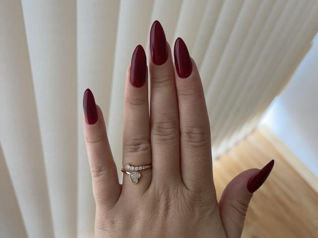 SugarrX's hand with long, pointed, red nails, wearing a gold ring with a small diamond, against a white, vertical, pleated backdrop.