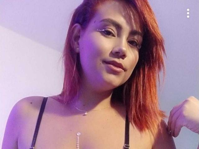 Sharon, con cabello rojo, top de tirantes negros, delineador de ojos y collar, sonriendo a la cámara, mostrando senos pequeños, piel clara y rostro lindo, con la mano cerca del hombro, luz púrpura..