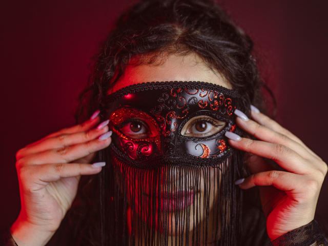 HelaViKy, con cabello oscuro y rizado, lleva una máscara negra ornamentada con detalles rojos y naranjas, un borde inferior fruncido y uñas moradas, sosteniendo la máscara frente a su rostro, con una luz roja en el fondo..
