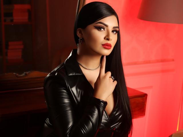 Devasha with long black hair, wearing a black leather jacket, red lipstick, and a silver necklace, pointing to her chin with her index finger. Red lighting in the background.