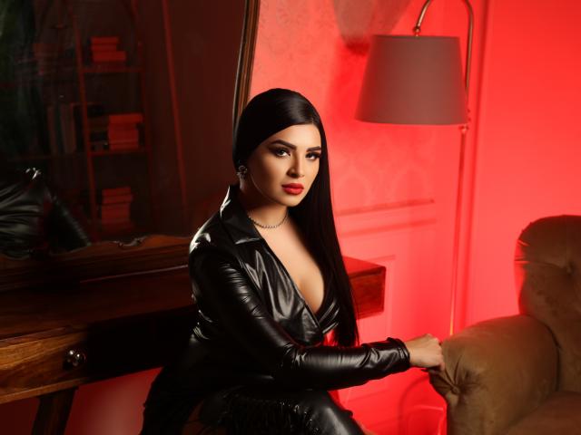Devasha with long black hair, wearing a black leather outfit, sitting on a couch, holding the armrest, red lighting, and a lamp in the background, looking seductive.