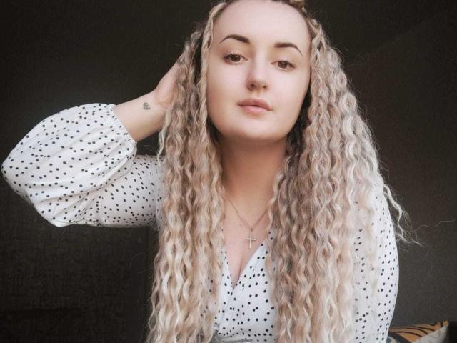 Jude Valenti, com longos cabelos loiros e cacheados, vestindo uma camisa branca com bolinhas, com a mão no cabelo, maquiagem discreta, um pequeno colar com uma cruz, expressão neutra, pele clara, uma pequena tatuagem no pulso..
