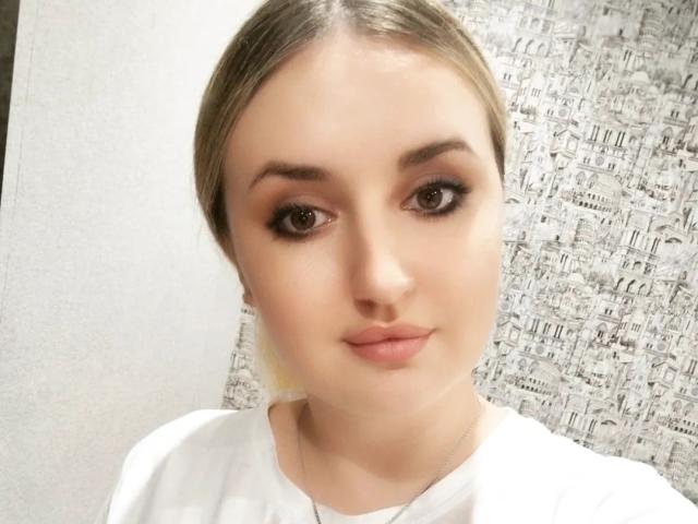JudeValenti has blonde hair, brown eyes, and is wearing a white shirt. She has a cute face with full lips and is looking at the camera with a neutral expression.