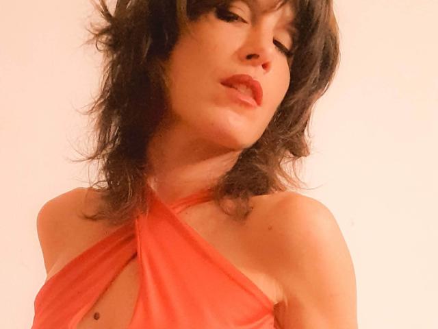 Aiyana with wavy black hair, red lipstick, and a red halter dress showing her cleavage and a small mole on her chest. She's looking down with a sultry expression.