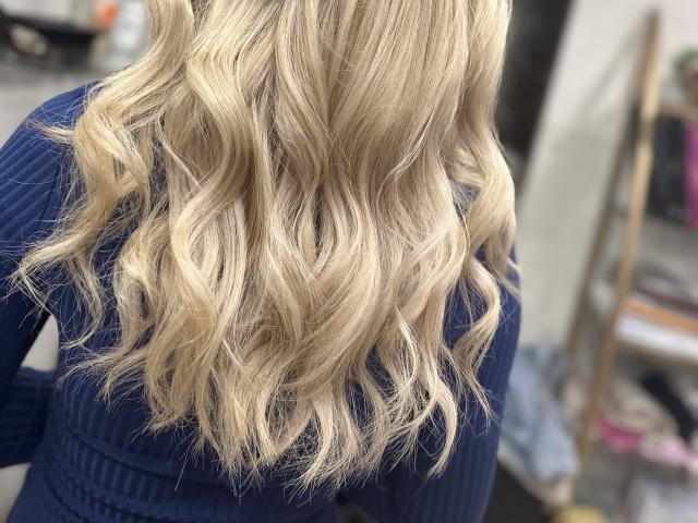 Les cheveux blonds, ondulés et mi-longs de SugarrX, coiffés en boucles douces et avec des reflets naturels, sont mis en valeur par un pull bleu en maille côtelée. Sans seins apparents, le visage est flou, l'attention est portée sur les cheveux..