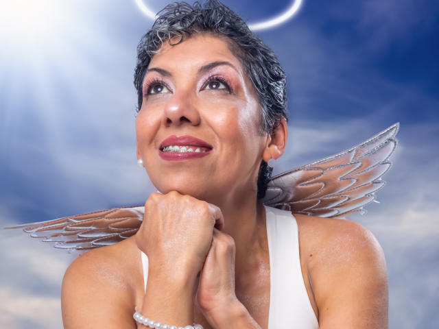 JuliaNewell has short curly black hair, a white halo, and transparent angel wings, smiling with her chin resting on her hand, wearing pink eyeshadow and a white sleeveless top.