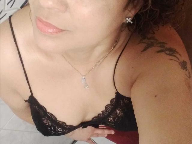 SolmaryHot with curly brown hair, glossy pink lips, black lace bra, key necklace, tattoo on right shoulder, holding bottom of bra, showing her tits and cleavage.