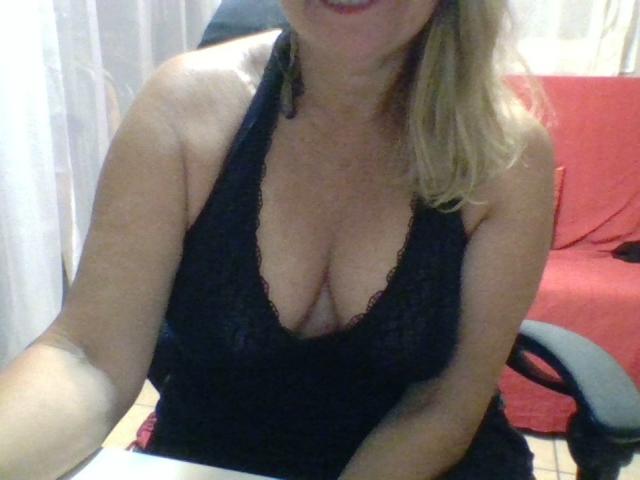 Coquine69x with blonde hair, wearing a black lace halter top, showing her big tits and cleavage, sitting in a chair, smiling, with a red couch in the background.
