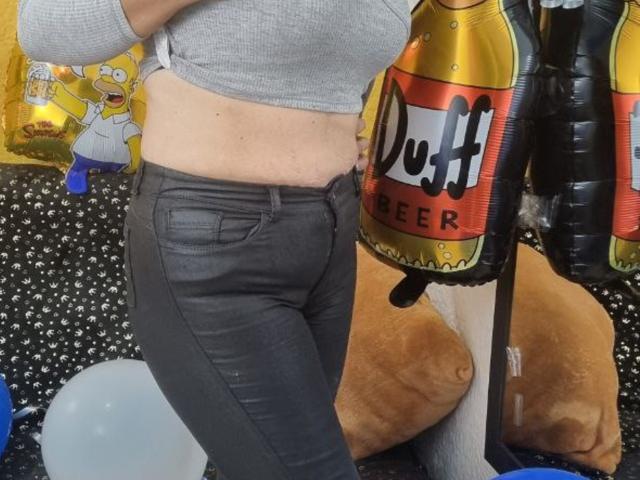 Le ventre légèrement apparent de CatalinaJH, ses pantalons en cuir noir, elle porte un haut court gris, avec un objet gonflable en forme de canette de bière sur la droite. Ses fesses sont visibles..