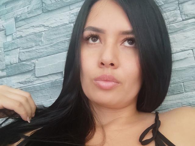 AshleyKatelyn has long black hair, brown eyes, and pink lips. She's making a pouty face, with one hand in her hair, wearing a black strappy top.
