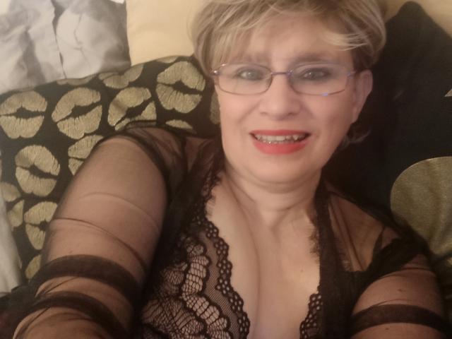 Samantha Mature hat kurze, blonde Haare, eine Brille und roten Lippenstift. Sie trägt eine schwarze Spitzenbluse, die ihre Brüste zeigt, und lächelt in die Kamera..