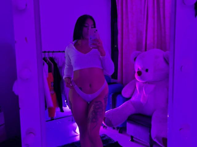 Amberv with dark hair, white crop top, and white panties, showing her curvy body and tattooed leg in a mirror selfie with purple lighting, white teddy bear in background.