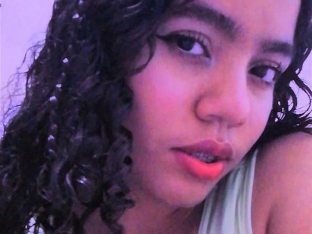 LarhaDani, con cabello negro rizado, ojos oscuros y lápiz labial rojo, mirando ligeramente hacia un lado con un leve puchero, vestida con una blusa verde claro, en un selfie con iluminación púrpura..