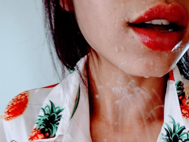 Les lèvres rouges d'Evelyn Mistri, légèrement entrouvertes, avec du sperme visible sur son menton et son cou, portant une chemise à motifs ananas, montrant une texture humide et brillante sur sa peau..