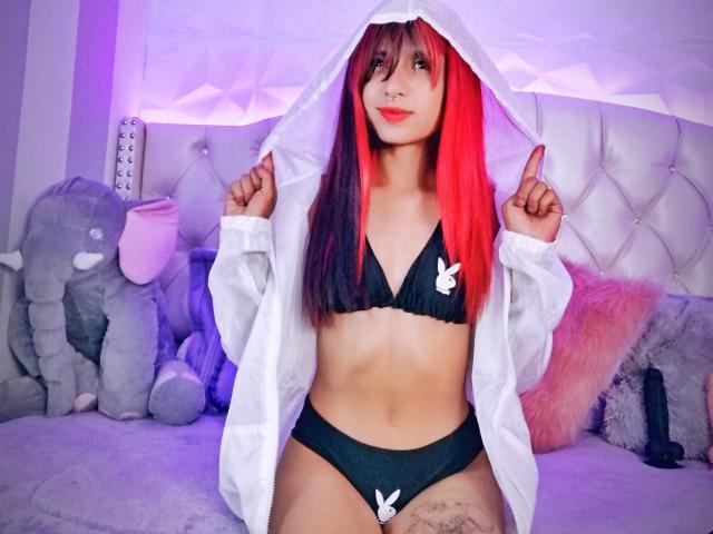 EvelynMistri with red and black hair, wearing a black bikini and white hoodie, pointing up with fingers, showing her tits, small ass, and tattoo on her right thigh.