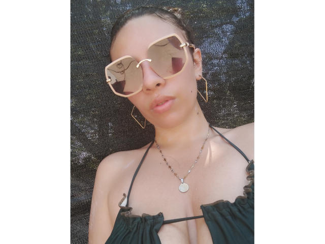 VioletaStone with tan skin, wearing large aviator sunglasses, gold hoop earrings, and a black ruffled bikini top, showing her tits. She's lying down, looking at the camera.