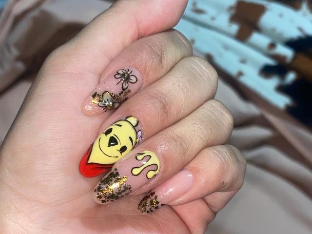 A mão de ShannelTaylor, com unhas sem esmalte, pontas com glitter e detalhes artísticos: um rosto amarelo com um "V" e um "Q" vermelhos, além de uma borboleta e uma flor na unha do dedo anelar..