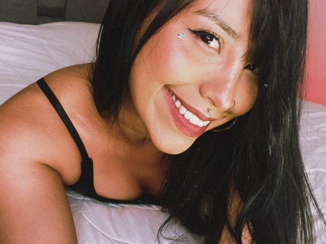 ElizabethMacy has long black hair, a nose ring, and a smile showing her white teeth. She's wearing a black strap top, lying on a bed with white sheets.