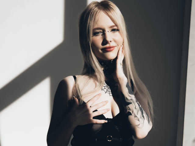LaneyvilI has blonde hair, blue eyes, and is smiling at the camera. She's wearing black lingerie, covering her tits with one hand, and has a tattoo on her arm.