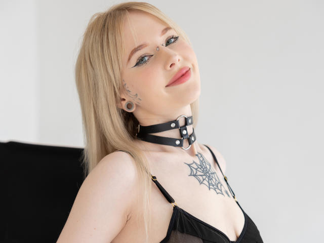 LaneyvilI bionda, con occhi azzurri, eyeliner nero e rossetto rosa, indossa una collana di pelle nera, ha un tatuaggio a forma di ragnatela sul petto e indossa biancheria intima nera, guarda verso l'alto con un leggero sorriso..