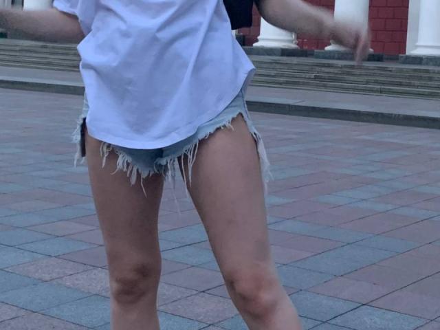 Tsereras weißes Hemd, die ausgefransten blauen Shorts, die ihr Gesäß und ihre Oberschenkel zeigen, mit leicht erhobenen Armen, stehend auf einem gefliesten Boden. Ihre Haut ist hell, und sie befindet sich im Freien..
