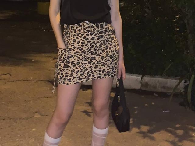 Tserera, com pele clara, vestindo uma blusa preta, uma saia com estampa de leopardo, meias brancas até o joelho e segurando uma bolsa preta, está em pé em um chão sujo e mal iluminado à noite..